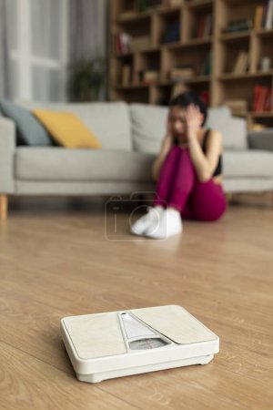 Photo for Weight-loss fail. Depressed korean lady crying, sitting near weight-scales after weighing herself, gaining weight, not losing excess kilograms on diet. Selective focus, shallow depth - Royalty Free Image