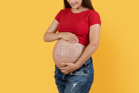 Foto de Pregnancy Concept. Cropped Shot Of Young Pregnant Woman Embracing Belly, Beautiful Smiling Female Expecting Baby Tenderly Touching Her Tummy While Standing Isolated Over Yellow Background In Studio - Imagen libre de derechos