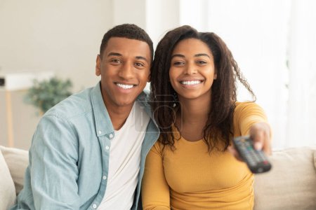 Photo for Smiling millennial african american husband hug his wife, switch channel with remote control, watch tv in spare time in light living room interior. Movie evening together at weekend, good news at home - Royalty Free Image