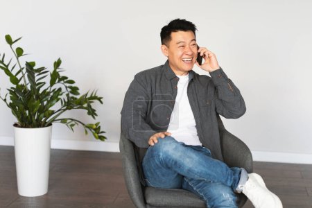 Photo for Cheerful middle aged asian man calling by phone, sitting in armchair in living room interior with white wall, copy space. Gadget for business communication, work remotely, talk with client at home - Royalty Free Image