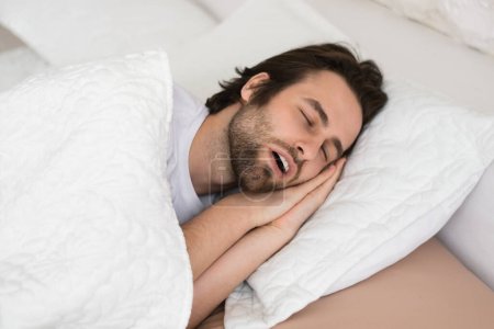 Tired young caucasian male with open mouth sleeping on white soft bed in bedroom in spare time, enjoy quiet alone, copy space, close up. Snoring, rest and relaxation at weekend, healthy sleep at night