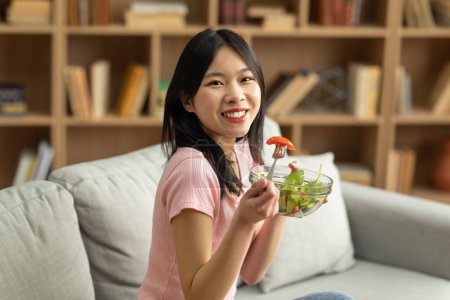 Téléchargez les photos : Healthy vegan diet concept. Happy asian lady sitting on sofa at home with bowl of fresh vegetable salad, eating tasty lunch. Young female enjoying her vegan meal, promoting clean living - en image libre de droit