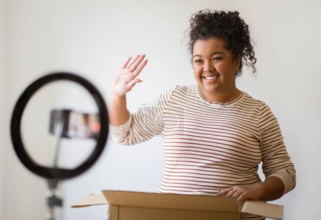Photo for Positive friendly attractive hispanic overweight millennial lady in casual outfit streaming while opening delivery, standing next to paper box, waving at smartphone and smiling, home interior - Royalty Free Image