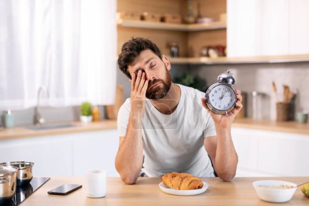 Foto de Unhappy tired sleepy millennial caucasian male with beard with alarm clock rubs his eyes at table, has breakfast alone in kitchen interior. Sleep deprivation, waking up early, overwork, time to work - Imagen libre de derechos