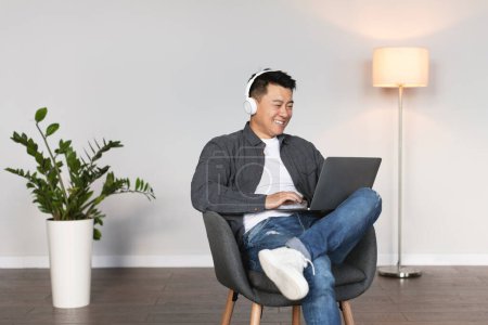 Photo for Glad adult chinese guy in wireless headphones listen to music and watch video on laptop in living room interior on white wall background. Rest with technology, business meeting, work remotely at home - Royalty Free Image