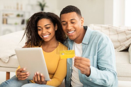 Téléchargez les photos : App for online shopping. Glad millennial african american guy hug lady, use tablet, credit card for order purchase, check banking in living room interior. Shopaholics enjoy sale and cashback at home - en image libre de droit