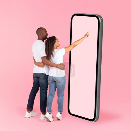 Foto de Rear back view of black spouses using big smartphone with blank screen, woman touching huge display panel, man hugging lady, standing on pink background, mock up - Imagen libre de derechos