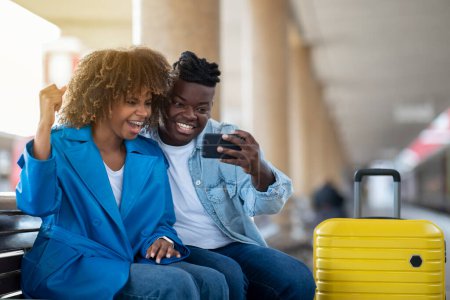 Foto de Online Win. Portrait Of Joyful Black Couple Celebrating Success With Smartphone While Sitting On Bench At Railway Station, Happy African American Man And Woman Using Mobile Phone While Waiting Train - Imagen libre de derechos