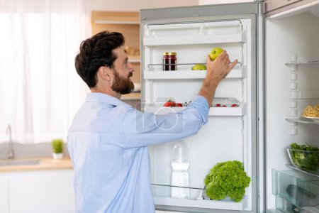 Photo for Happy hungry millennial caucasian bearded guy opens refrigerator door and takes green apple for cooking food in kitchen interior, copy space. Diet, vegetarianism, health care and lifestyle at home - Royalty Free Image