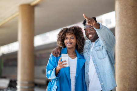 Photo for Happy Black Couple Waiting For Train At Railway Station Together, Cheerful Young African American Spouses Standing On Platform, Woman Holding Smartphone And Man Pointing Away, Free Space - Royalty Free Image