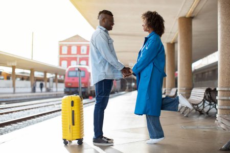 Téléchargez les photos : Romantic Black Couple Standing On Platform At Railway Station And Holding Hands, Loving Young African American Man And Woman Having Tender Moments While Waiting For A Train Arrival, Side View - en image libre de droit