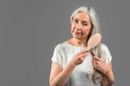 Téléchargez les photos : Serious senior caucasian female combing her gray hair with comb, isolated on gray background, studio, copy space. Beauty care at home daily procedure, treatments and problems with hair, ad and offer - en image libre de droit