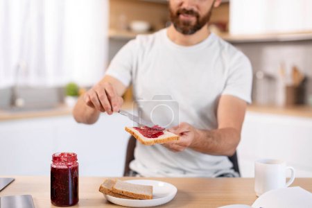 Téléchargez les photos : Smiling adult caucasian guy with beard making sandwich with jam at table, enjoy weekend in kitchen interior, cropped, close up. Breakfast alone at home, good morning and healthy food at spare time - en image libre de droit