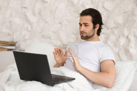 Téléchargez les photos : Sad serious young caucasian guy sit on bed, looks at computer, has video call, makes gesture calm down with his hands, psychological help in bedroom. Communication remotely, tech for meeting at home - en image libre de droit
