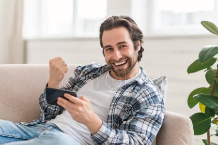 Téléchargez les photos : Satisfied young caucasian guy on sofa with smartphone making victory sign with hand, enjoy success in win in online game in room interior. Blog, new app, great ad and offer, huge sale and good news - en image libre de droit