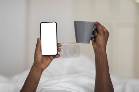 Téléchargez les photos : Black Man Holding Blank Smartphone And Cup Of Coffee While Resting In Bed At Home, Unrecognizable African American Guy Using Modern Gadget With Copy Space For Website Or Mobile App Design, Mockup - en image libre de droit