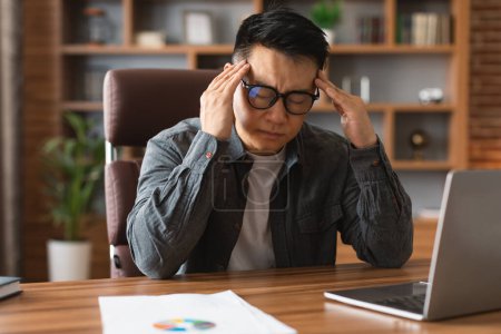 Téléchargez les photos : Despaired unhappy adult korean guy in glasses works on laptop, presses hands to temples, suffers from problems in business and headache in office interior. Migraine, overwork, illness and deadline - en image libre de droit