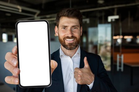 Téléchargez les photos : Happy caucasian male entrepreneur in suit showing smartphone with empty screen and gesturing thumb up in office. Businessman advising app for trade, offer and business recommendation - en image libre de droit