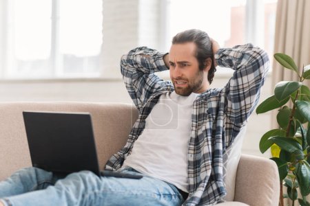 Foto de Cheerful young caucasian guy sits on sofa with computer, watch video, resting, enjoy spare time alone in living room interior. Blog, relax with device, ad and offer, video call and social distance - Imagen libre de derechos