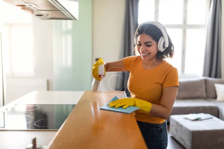 Foto de Smiling Young Arab Female In Wireless Headphones Making Cleaning At Home, Happy Middle Eastern Woman Using Detergent Sprayer And Rag While Washing Kitchen Counter From Dust, Copy Space - Imagen libre de derechos