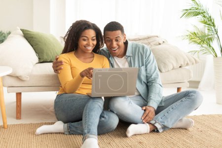 Téléchargez les photos : Happy millennial african american husband hug wife, watch video on laptop, have online call, sit on floor in living room interior. Meeting remote, technology for shopping, surfing in internet at home - en image libre de droit