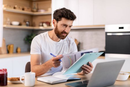 Téléchargez les photos : Busy smiling millennial caucasian businessman with computer analyzes data, works with charts in kitchen interior. Good morning, business and freelance at home, startup idea and financial accounting - en image libre de droit