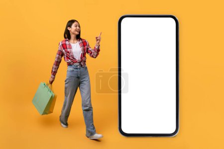 Téléchargez les photos : App for online store. Full body length of asian lady pointing at big smartphone with blank white screen, walking with shopping bags on yellow background, mock up banner - en image libre de droit