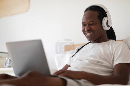 Photo for Online Education. Young Black Man Study With Laptop While Sitting In Bed At Home, Smiling African American Guy Wearing Wireless Headphones Using Computer For Distance Learning, Closeup Shot - Royalty Free Image