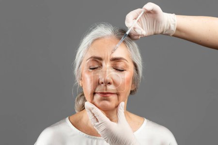 Foto de Serious old caucasian lady with closed eyes on beauty procedure, cosmetologist hands in gloves make botox injection, isolated on gray background, studio. Anti-aging skin care, professional treatment - Imagen libre de derechos