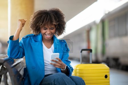 Fröhliche schwarze Frau feiert Erfolg mit Smartphone, während sie auf Bank am Bahnhof sitzt, fröhliche Afroamerikanerin spielt Online-Spiele, hat Spaß beim Warten auf Zug, freier Platz