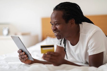 Téléchargez les photos : Online Purchasing. Cheerful Black Man Using Digital Tablet And Credit Card While Relaxing In Bed At Home, Happy African American Guy Shopping In Internet While Resting In Bedroom, Closeup - en image libre de droit