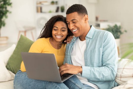 Foto de Technology at home. Glad millennial african american wife hugging husband, looking at laptop, have video call in living room interior. New normal for communication, search, ad and offer due covid-19 - Imagen libre de derechos