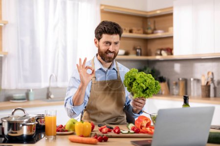 Foto de Glad millennial caucasian male cooking eat show salad and ok hand sign at table with vegetables and computer in kitchen interior. Chef recommendation and advice, new recipe, food blog and video call - Imagen libre de derechos