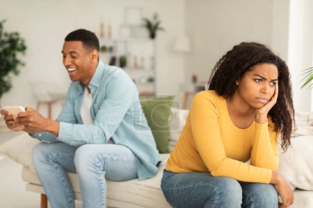 Photo for Angry offended millennial african american lady sad at guy with phone, suffer from addiction to games and social networks in living room interior. Emotions, relationships problems and crisis at home - Royalty Free Image