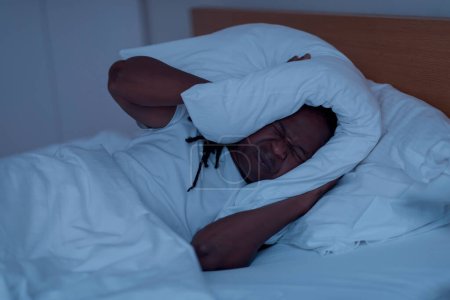 Téléchargez les photos : Sleep Disorders. Annoyed Black Man Lying In Bed And Covering Head With Pillow, Irritated Young African American Guy Suffering Insomnia While Resting In bedroom During Night Time, Closeup - en image libre de droit