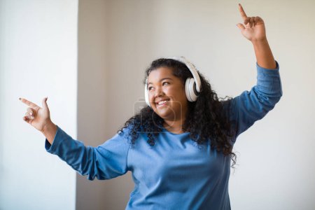 Foto de Best music. Nice positive happy young plus size woman in homewear dancing to her favourite song while having fun at home, looking through the window and smiling, using wireless headphones - Imagen libre de derechos