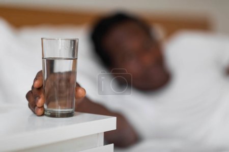 Téléchargez les photos : Thirsty Black Man Taking Glass Of Water From Bedside Table In Bedroom, Young African American Male Reaching Hand To Healthy Drink After Waking Up In The Morning, Selective Focus With Free Space - en image libre de droit