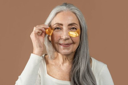 Foto de Smiling old caucasian female with natural beauty applies patches under eyes, isolated on brown background, close up, studio. Anti-aging procedures, fight against wrinkles, beauty care and renovation - Imagen libre de derechos