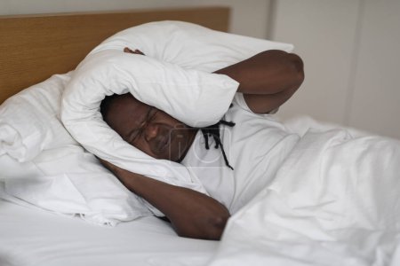 Photo for Irritated Black Man Lying In Bed And Covering Ears With Pillow, Annoyed Young African American Male Suffering Insomnia, Frowning Guy Emotionally Reacting To Loud Noise From Neighbours, Copy Space - Royalty Free Image