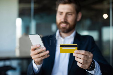 Téléchargez les photos : Male entrepreneur sitting at workdesk, using smartphone and holding credit card, shopping on Internet while working at office. Financial, banking, shopping mobile app - en image libre de droit