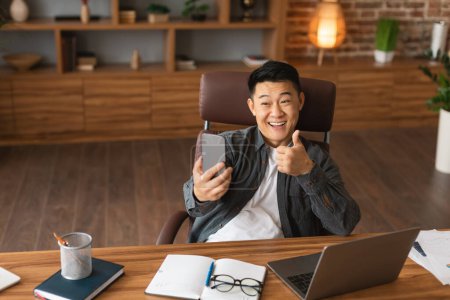 Photo for Cheerful mature asian man has video call, showing thumb up gesture in webcam, approve great deal at workplace with laptop in office interior. Meeting, recommendation and advice online startup business - Royalty Free Image