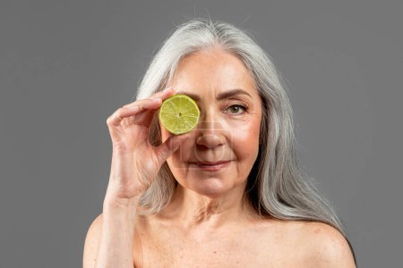 Foto de Serious old european female with gray hair puts lemon to eye, isolated on gray background, studio. Beauty care, anti-aging treatment, fight with wrinkle, rejuvenation and moistening skin, ad and offer - Imagen libre de derechos