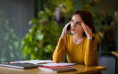 Photo for Anxious unhappy attractive young woman have phone conversation, female freelancer sitting at table with notepads and laptop on, working at coffee shop, looking at copy space, touching head - Royalty Free Image