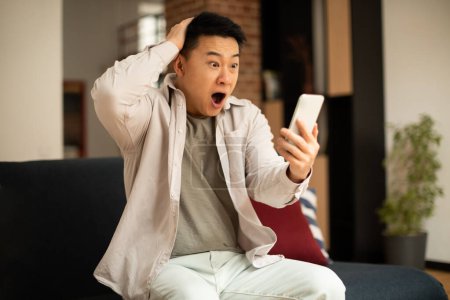 Foto de Oh no. Shocked mature asian man reading bad message on smartphone while sitting on sofa at home, free space. Male emotionally reacting to unpleasant news - Imagen libre de derechos