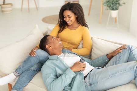 Téléchargez les photos : Cheerful millennial african american couple with remote control enjoy free time and tender moment, relaxing, lying on sofa in light living room interior. Movie night at home, love, romance and tech - en image libre de droit