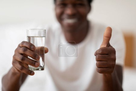 Téléchargez les photos : Joyful African American Man Showing Glass Of Water And Gesturing Thumb Up At Camera, Smiling Black Guy Recommending Healthy Refreshing Mineral Drink While Relaxing At Home, Selective Focus - en image libre de droit