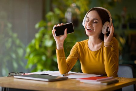 Foto de Joyful pretty young woman freelancer working at cafe, sitting at table with laptop, notepads on, singing with closed eyes, using phone with empty black screen, wireless headphones, copy space, mockup - Imagen libre de derechos