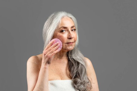 Foto de Serious old caucasian lady in towel cleans face with lifting sponge, isolated on gray background, studio. Beauty care, removing make up, anti-aging treatment at home, spa procedures for perfect skin - Imagen libre de derechos