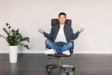 Téléchargez les photos : Serious calm adult asian male with closed eyes sits in armchair, meditates, rests from work, enjoys silence and free time in living room interior with white wall. Relax at home, break, lifestyle - en image libre de droit