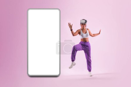 Photo for Dance moves in metaverse. Young woman dancing in virtual reality headset near huge smartphone with blank screen, pink background, mockup. Black lady having fun while exploring 3D technology. - Royalty Free Image